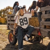 Load image into Gallery viewer, A woman sitting on a wooden farm trailer holds hands with a man coming out of the trailer, with other people and hay bales in the background. She is wearing a cream graphic t-shirt with '89' printed on the front, dark wash jeans, a plaid shirt tied around her waist, and a black baseball cap.