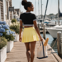 Load image into Gallery viewer, A woman walking on a wooden dock by the water, wearing a black short-sleeve crop top with white stripes and a bright yellow pleated mini skirt, carrying a canvas tote bag with a loaf of bread.