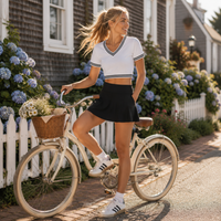 Load image into Gallery viewer, A woman stands on a vintage cream bicycle in front of a white picket fence and house, wearing a white cropped V-neck T-shirt with dark trim, a black mini skirt, and white sneakers.