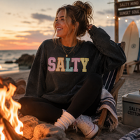 Load image into Gallery viewer, A young woman is on the beach near a campfire wearing Converse sneakers, leggings and a dark gray or faced black sweatshirt that says in faux embroidery, 'Salty" in pastel letters.