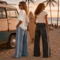 Load image into Gallery viewer, Two women standing on a beach at sunset, viewed from the back. One wears a white graphic T-shirt and light blue wide-leg jeans with white side stripes, and the other wears a white ringer T-shirt and dark gray wide-leg jeans with white side stripes.