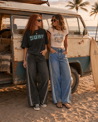 Load image into Gallery viewer, Two women standing on a sandy beach next to a vintage cream and blue van, wearing oversized streetwear-style outfits. One wears a dark charcoal t-shirt with 'SURF' in teal and dark wide-leg jeans with white side stripes.