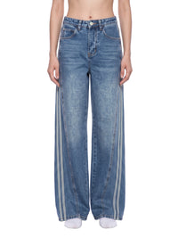 Load image into Gallery viewer, A model is wearing a pair of wide-leg blue denim jeans with three vertical white stripes down the outer side of each leg, shown against a plain white background.