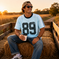 Load image into Gallery viewer, A man wearing a light blue t-shirt with a large black '89' graphic sits on a hay bale in a wooden trailer during golden hour. He is wearing blue jeans, white sneakers, sunglasses, and a tan cap, holding a coffee cup.