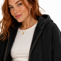 Load image into Gallery viewer, A close-up of a woman with red hair wearing a black hooded zip-up jacket over a white ribbed top, with a gold pendant necklace visible against a plain white background.